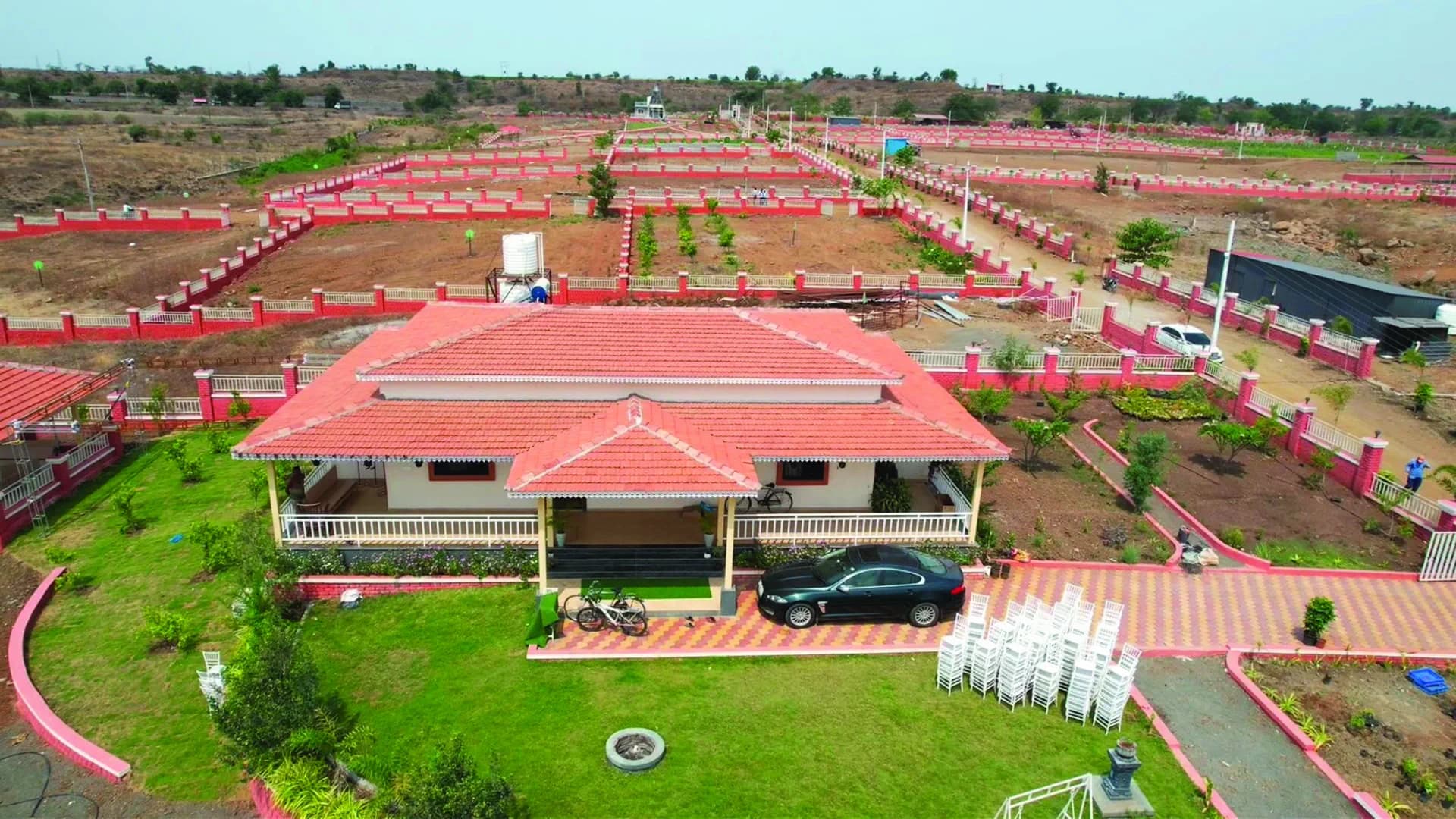 Aerial photo of Isha Farms — a completed farmhouse with a red-tile roof amid demarcated plots with pink boundary walls